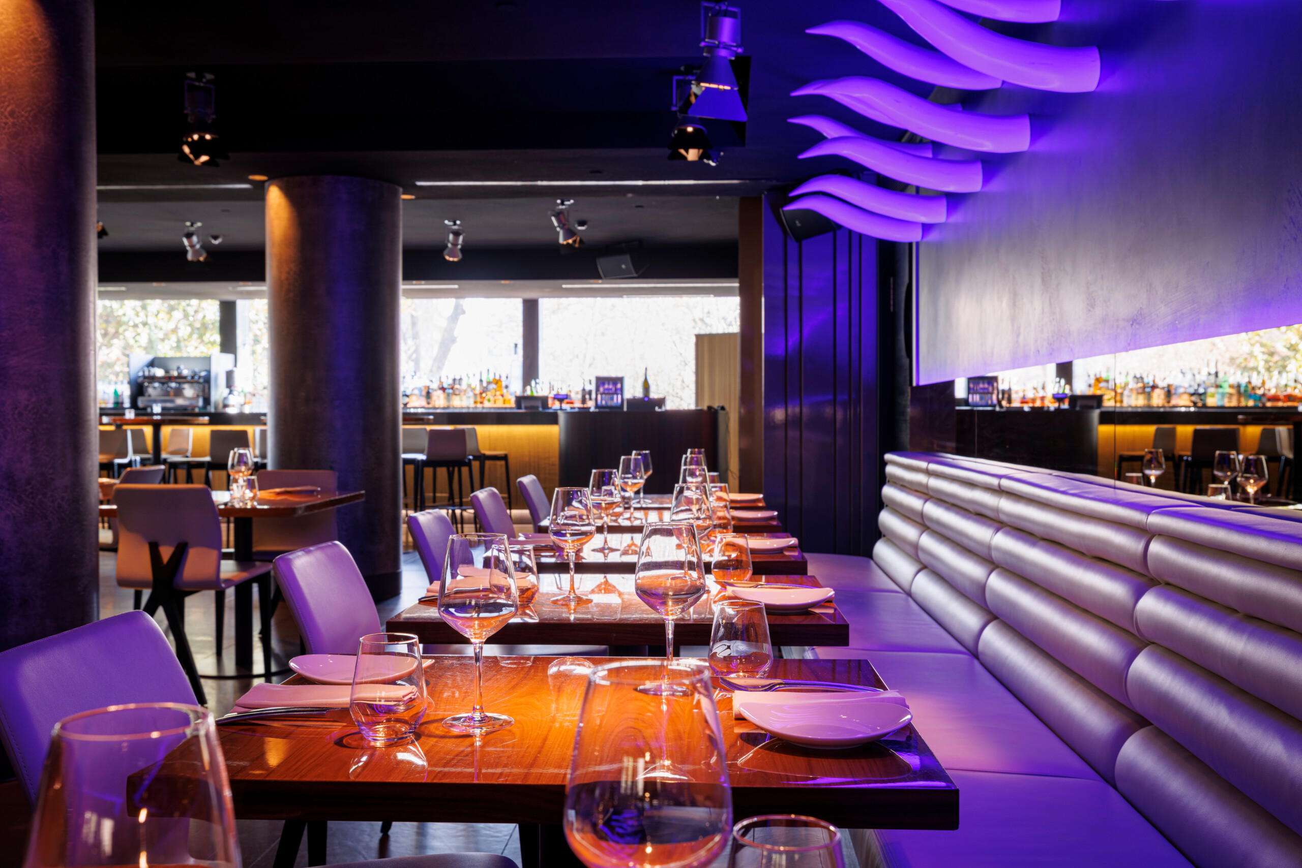Interior of a restaurant featuring dark wooden tables set with glassware and plates, surrounded by white chairs and a long, tufted banquette along one wall. The ceiling has dramatic, sculptural wave like panels illuminated by purple accent lighting