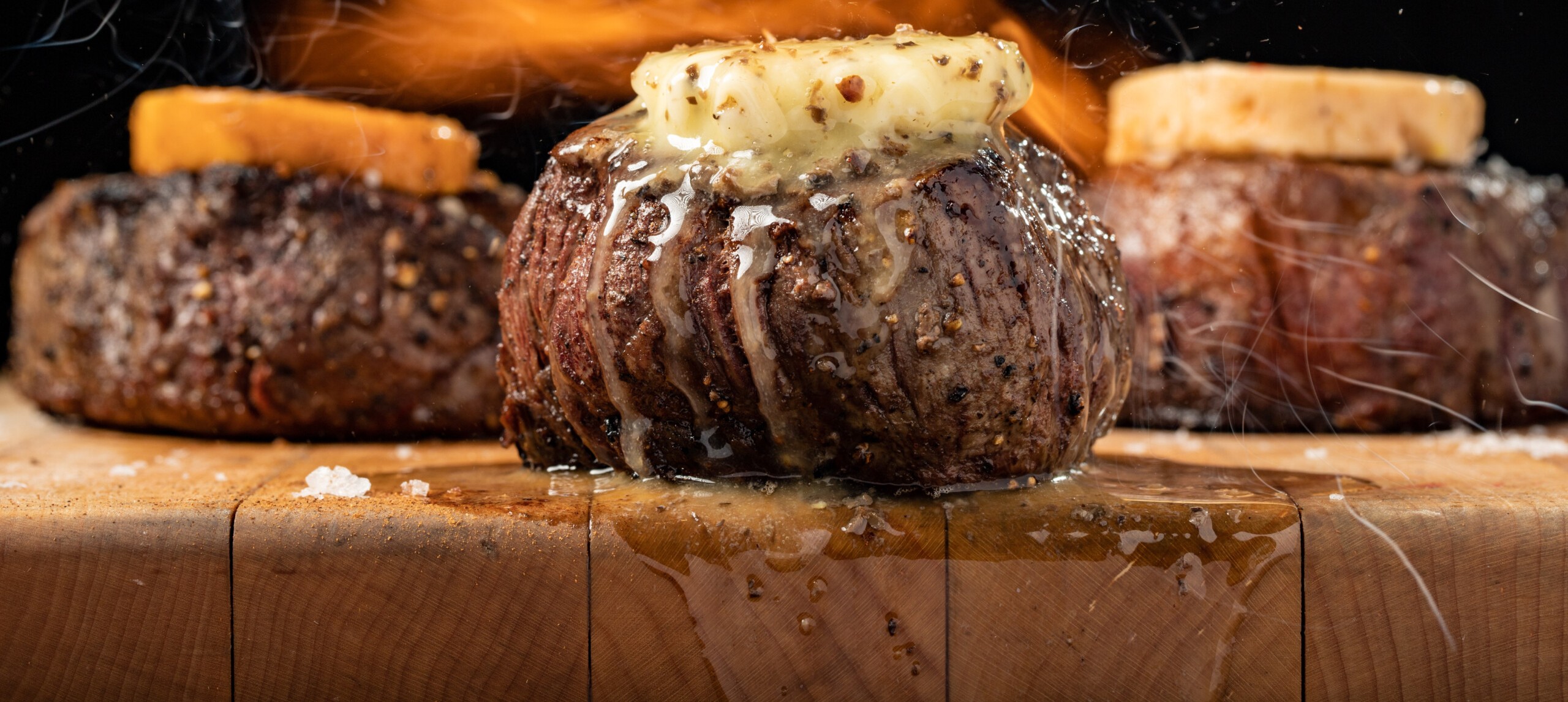 A trio of juicy steaks on a wooden chopping board, topped with butter that is dripping down the steaks