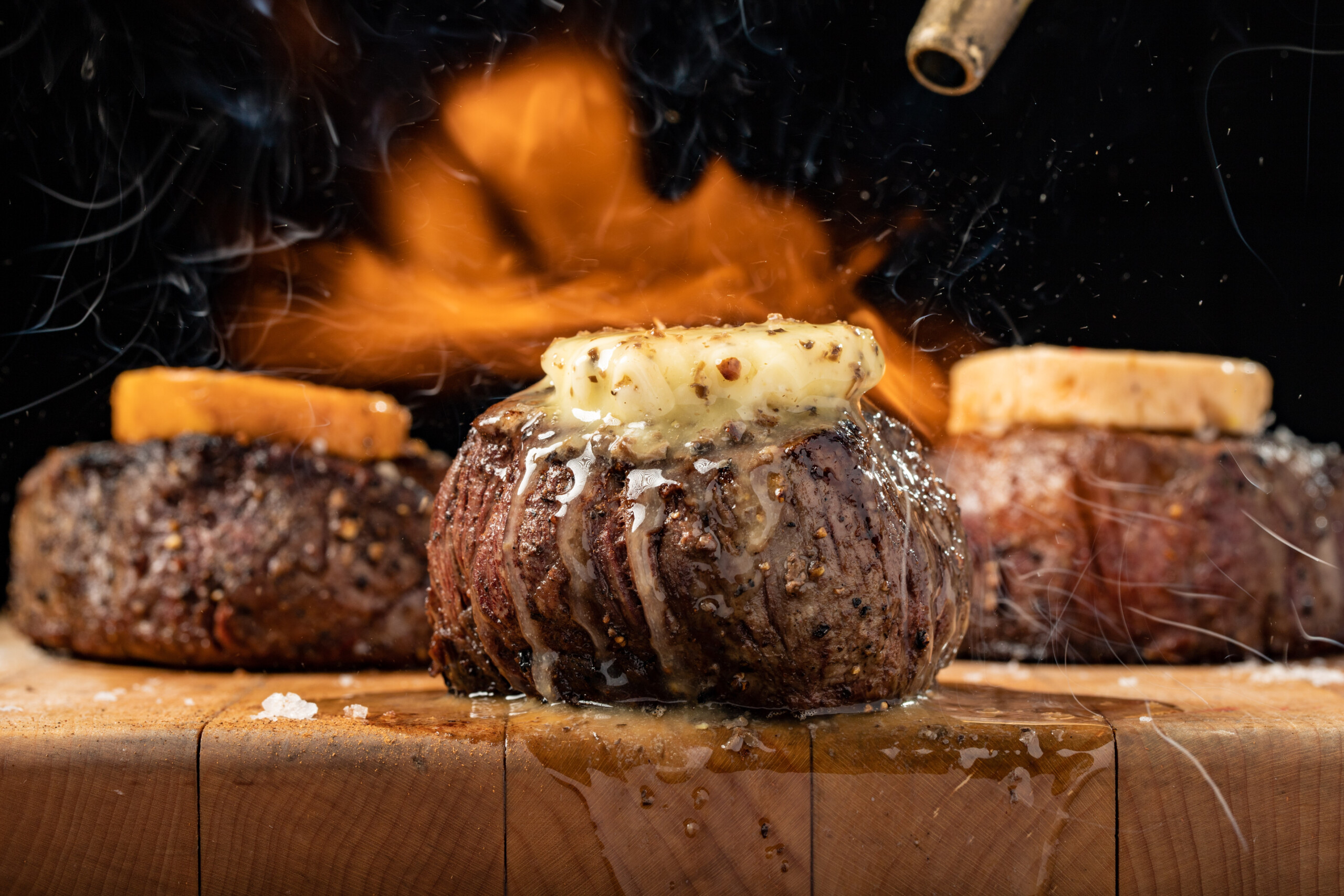 steak with cheese or butter melting on top, other steaks or burgers and a flame in the background