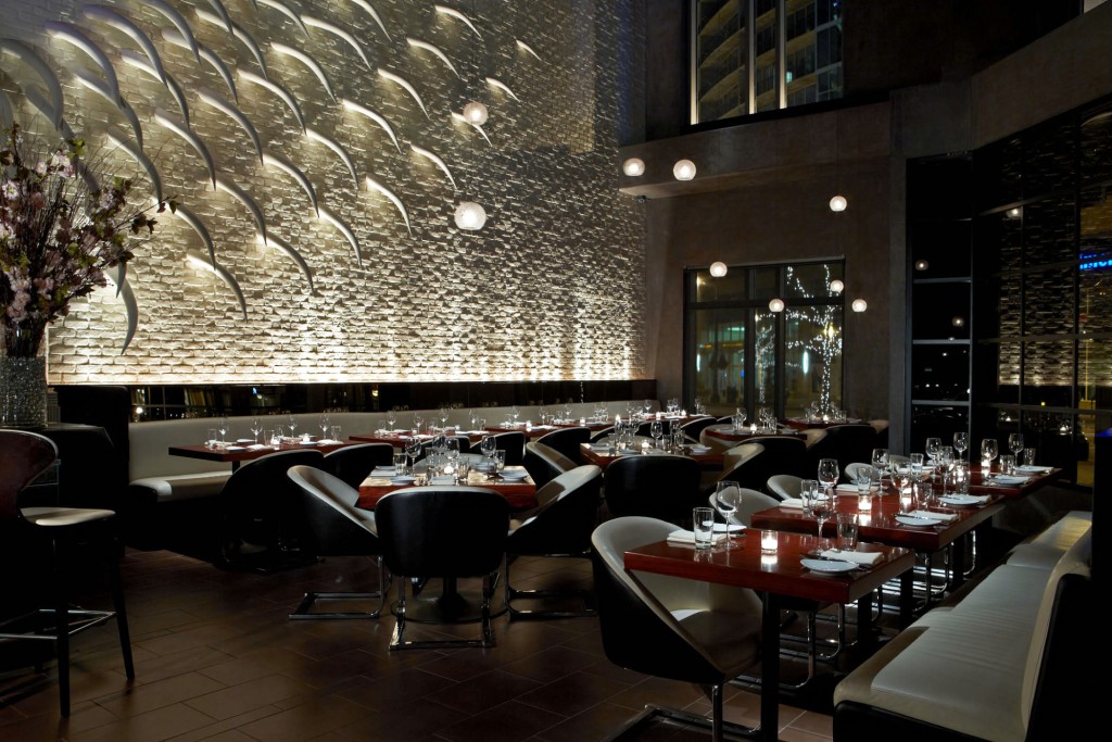 Interior of a restaurant with a textured back wall featuring a repeating pattern of white. The dining area includes dark wooden tables set with glasses and plates, surrounded by a mix of white chairs and curved white banquettes.
