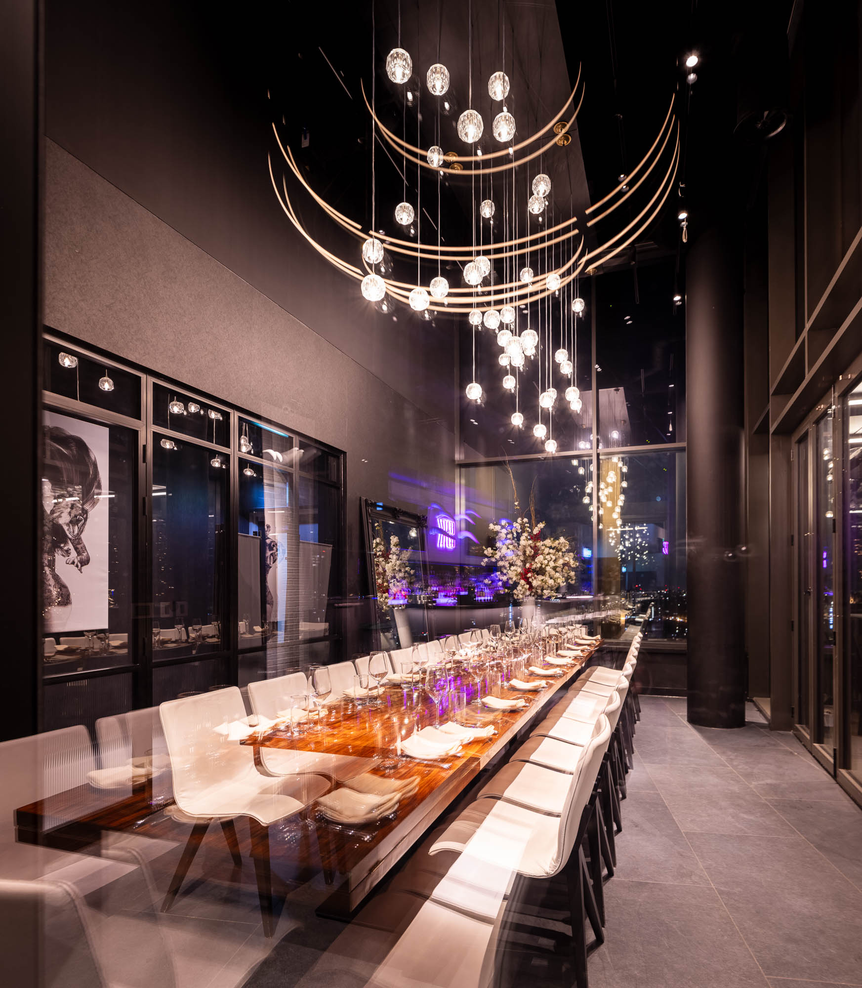 Interior of STK Stratford; a long row of tables and white seats in a soothingly lit room with high ceilings, chandeliers, and hanging artwork.