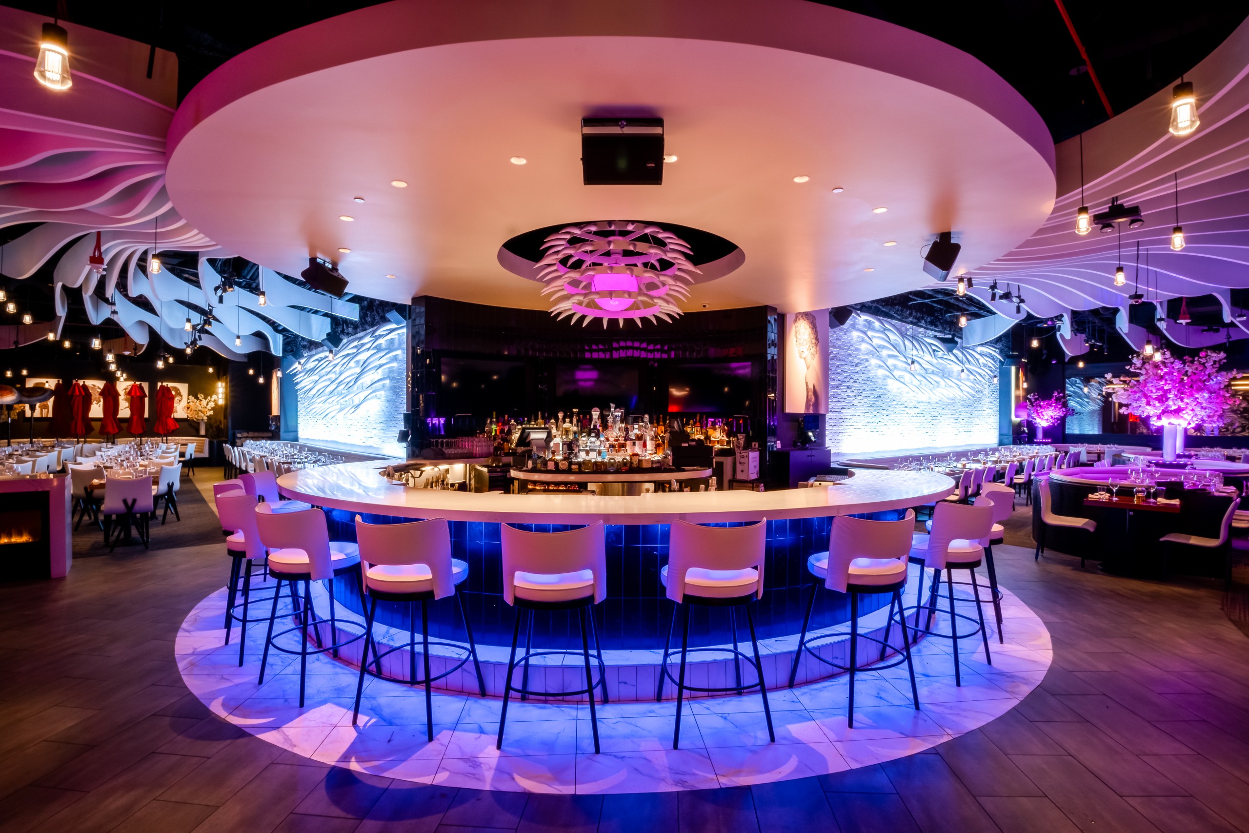 Interior of STK San Francisco bar and lounge with a large circular bar at the centre, surrounded by white barstools. The ceiling features a large, round architectural detail and the space is illuminated by purple and blue accent lights.