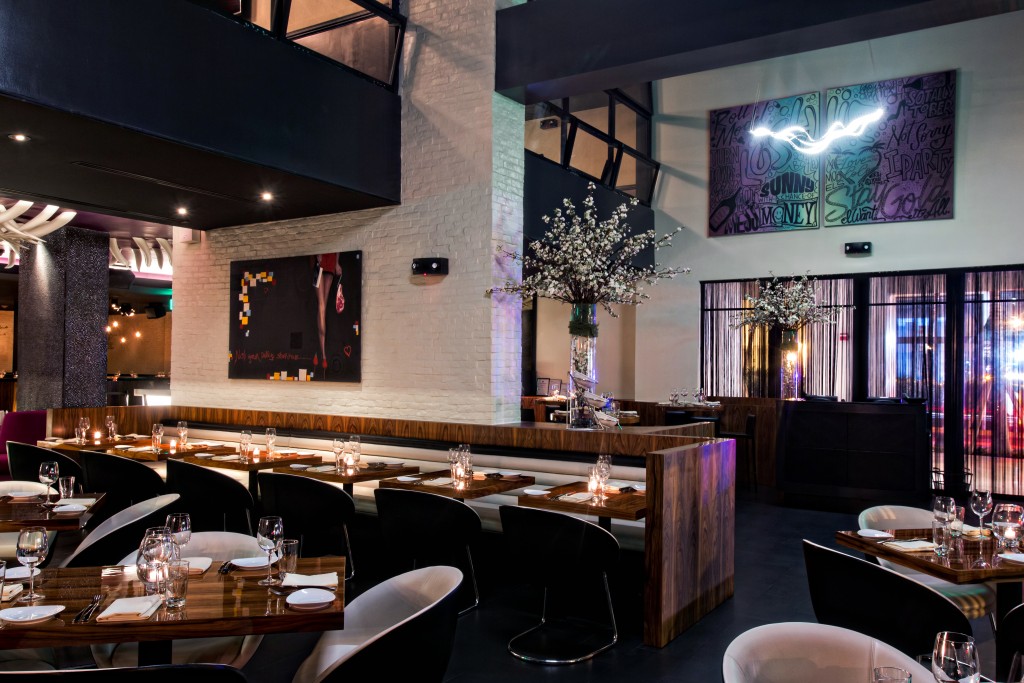 Interior of STK South Beach featuring dark wooden tables, dark chairs and ceiling, highlighted by a sculptural light fixture with looping neon like white tubing in the background. Tables are set with glassware and napkins, surrounded by contemporary white chairs with black legs.