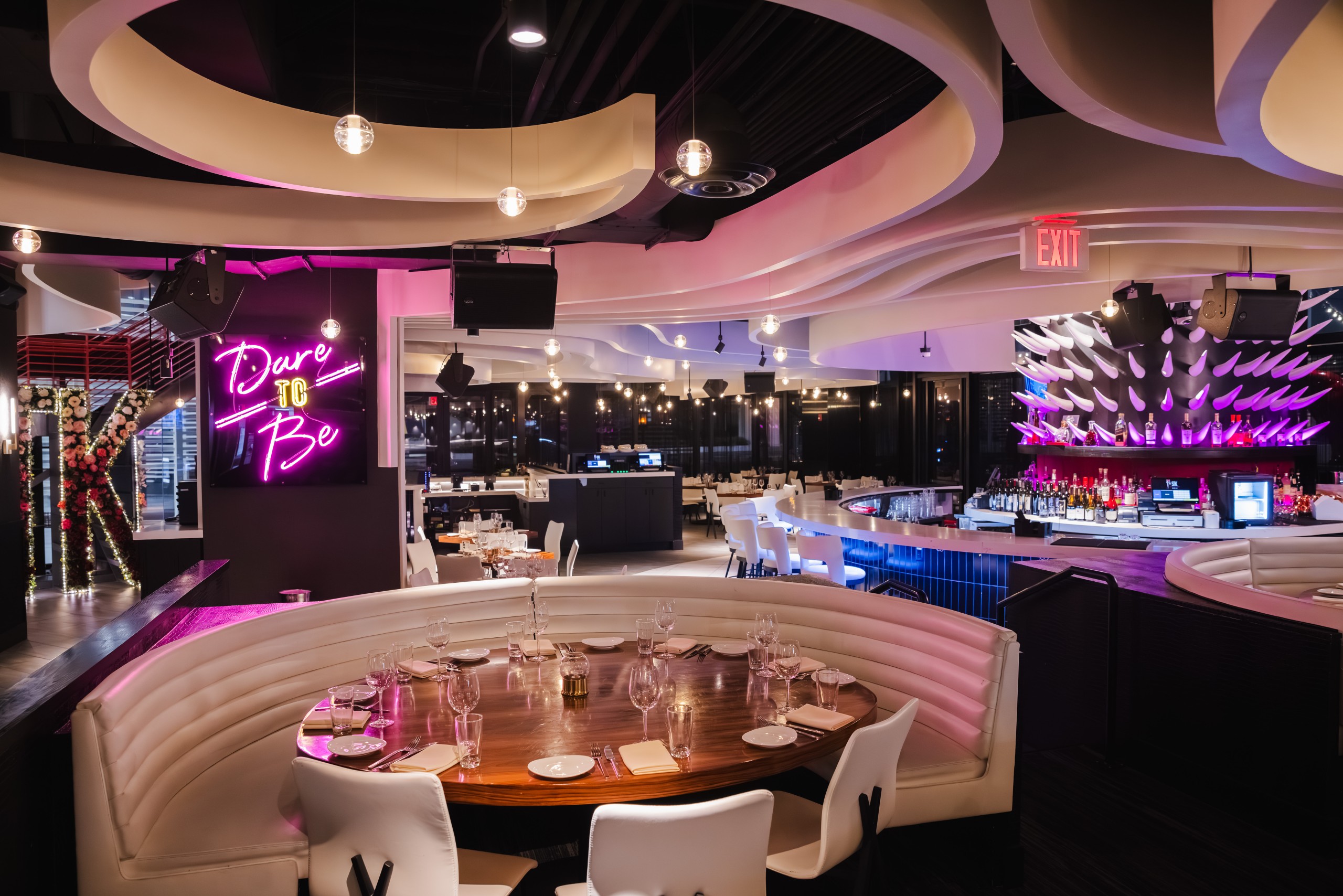 Interior of STK Charlotte restaurant featuring curved white banquette seating around a dark wooden table set with plates, glasses, and napkins. Overhead, the ceiling displays swirling white panels with built-in lighting. A neon pink sign reading 