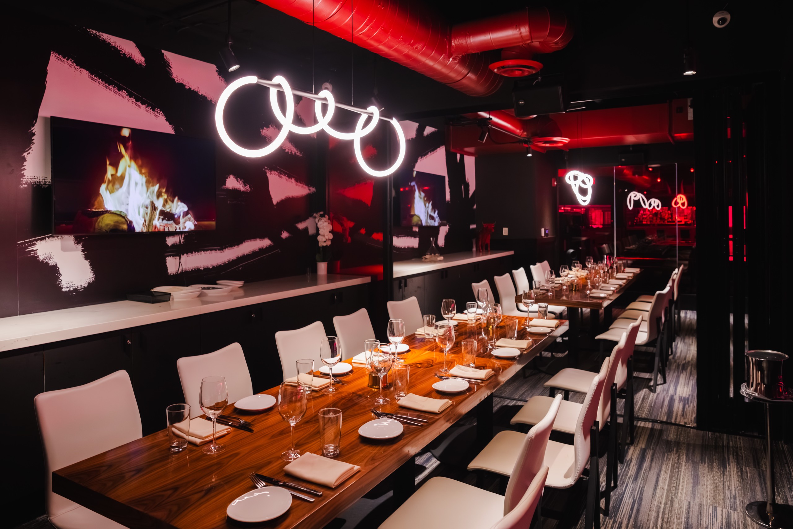 Interior of restaurant featuring a long wooden dining table set for service, surrounded by white chairs. The walls and ceiling are dark, accented by bold red lighting and a circular neon light fixtures hang above the table.