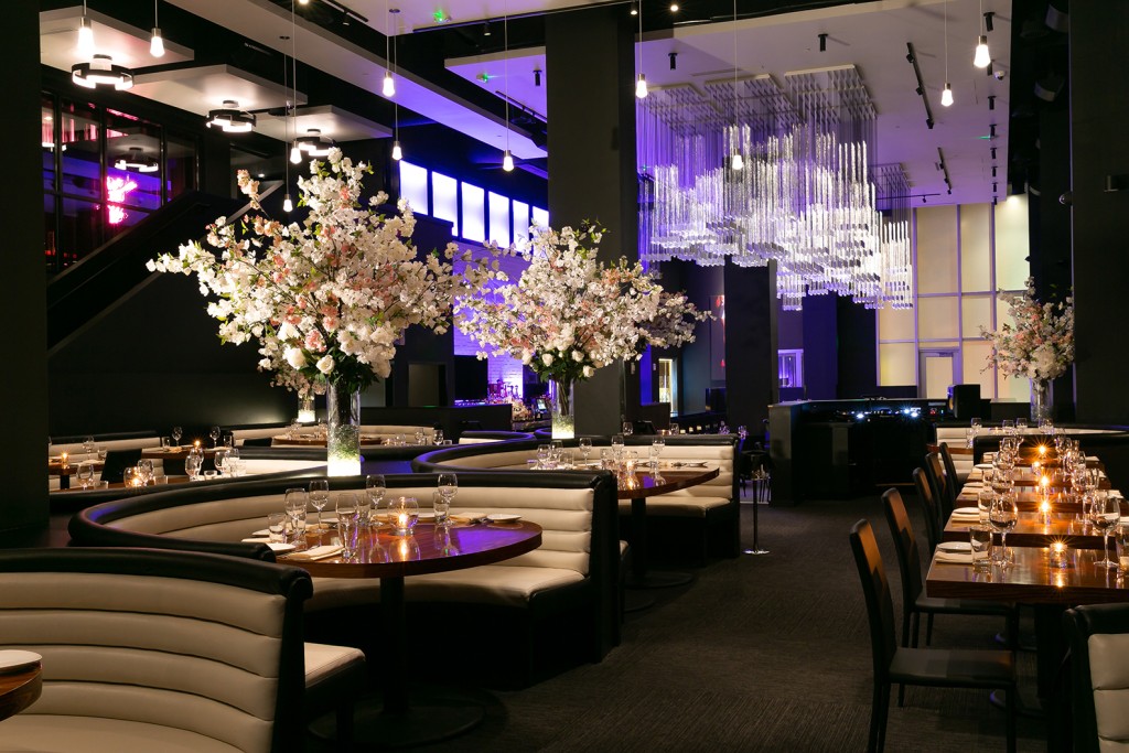 Interior of STK Nashville featuring curved white booth seating arranged in semi circles around round wooden tables set with glassware and plates. Tall, sculptural floral arrangements serve as striking centrepieces.