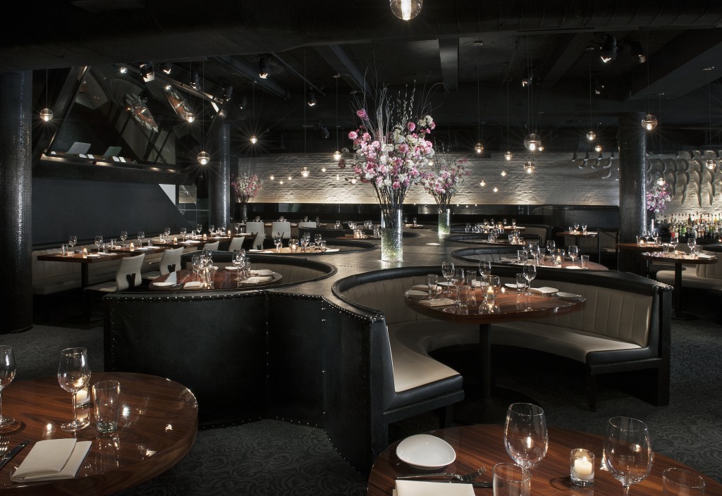 Interior of STK NYC Downtown featuring curved booth seating upholstered in dark material. The booths are arranged in a semi-circular pattern around round wooden tables, each set for service. At the centre of the space, there are tall floral arrangement.