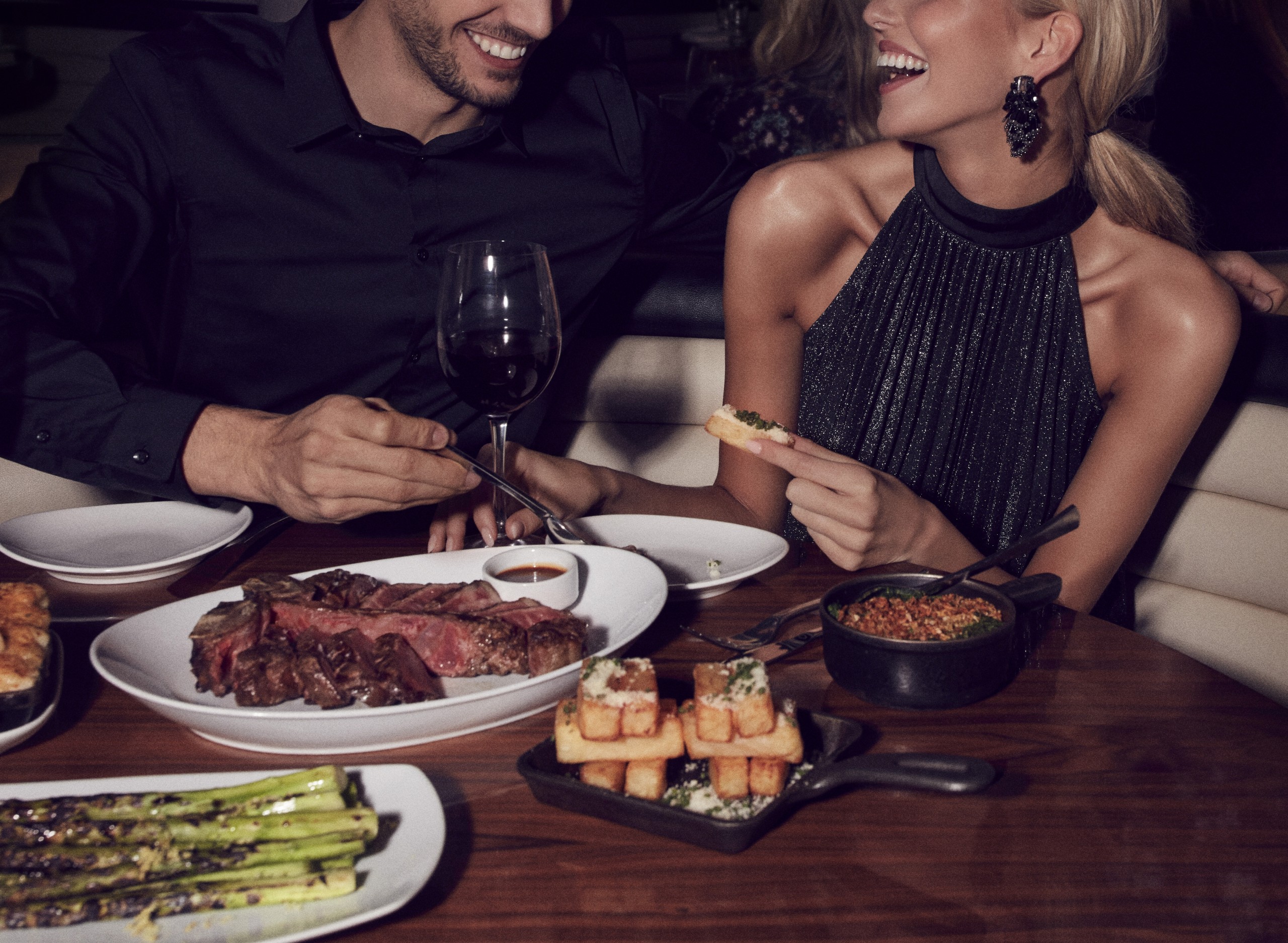 A man and a woman sitting in a booth having a nice time. On their table is a thick juicy steak, thick cut potato and asparagus
