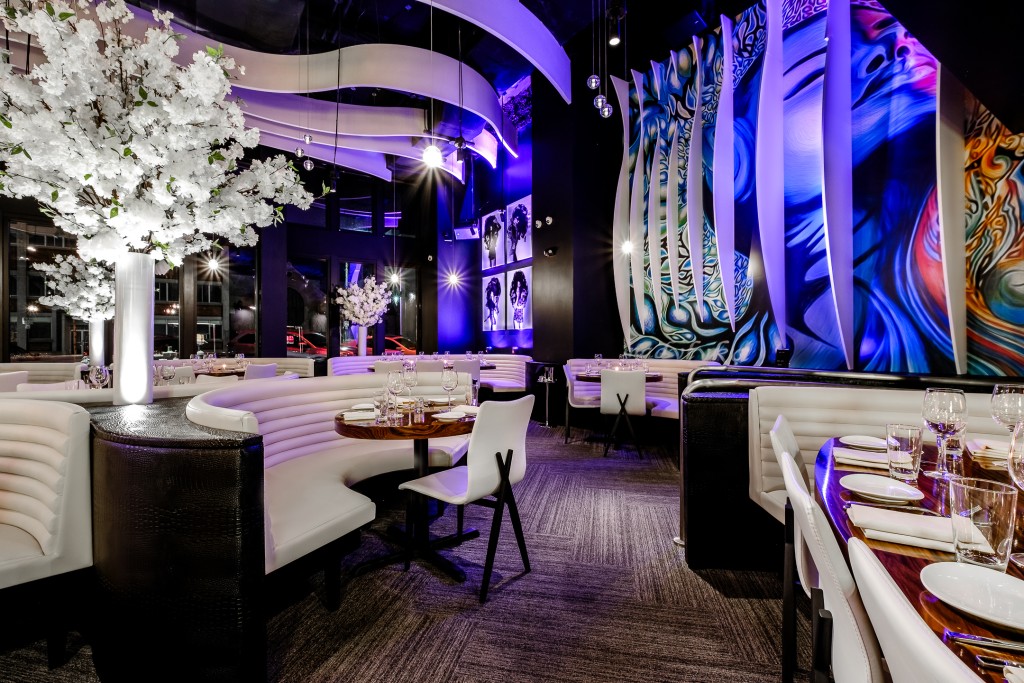 Interior of STK Salt Lake City restaurant featuring curved white banquettes and white chairs surrounding dark wooden tables, each set with plates, glasses, and napkins. White floral arrangements in tall vases are in between the booths. The ceiling has wavy white panels with purple accent lighting.